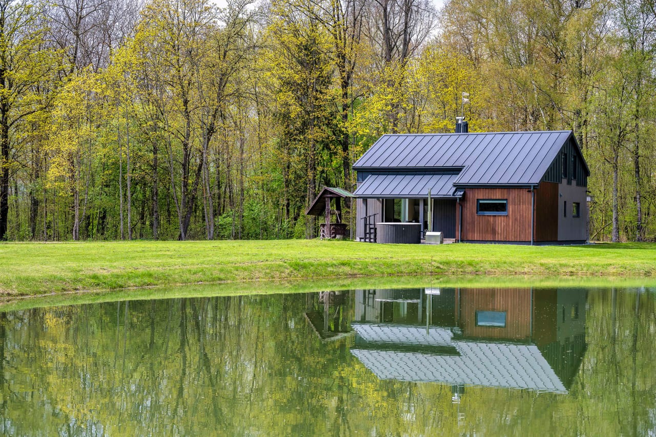 Vila Valentino cabin reflected in a still pond, with a wooden well beside the porch and spring forest behind